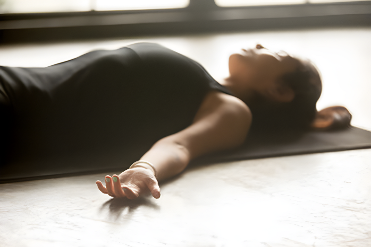 Young sporty woman practicing yoga at home, lying in Savasana exercise, Corpse pose, working out, wearing black sportswear, indoor closeup, studio floor background. Wellness concept